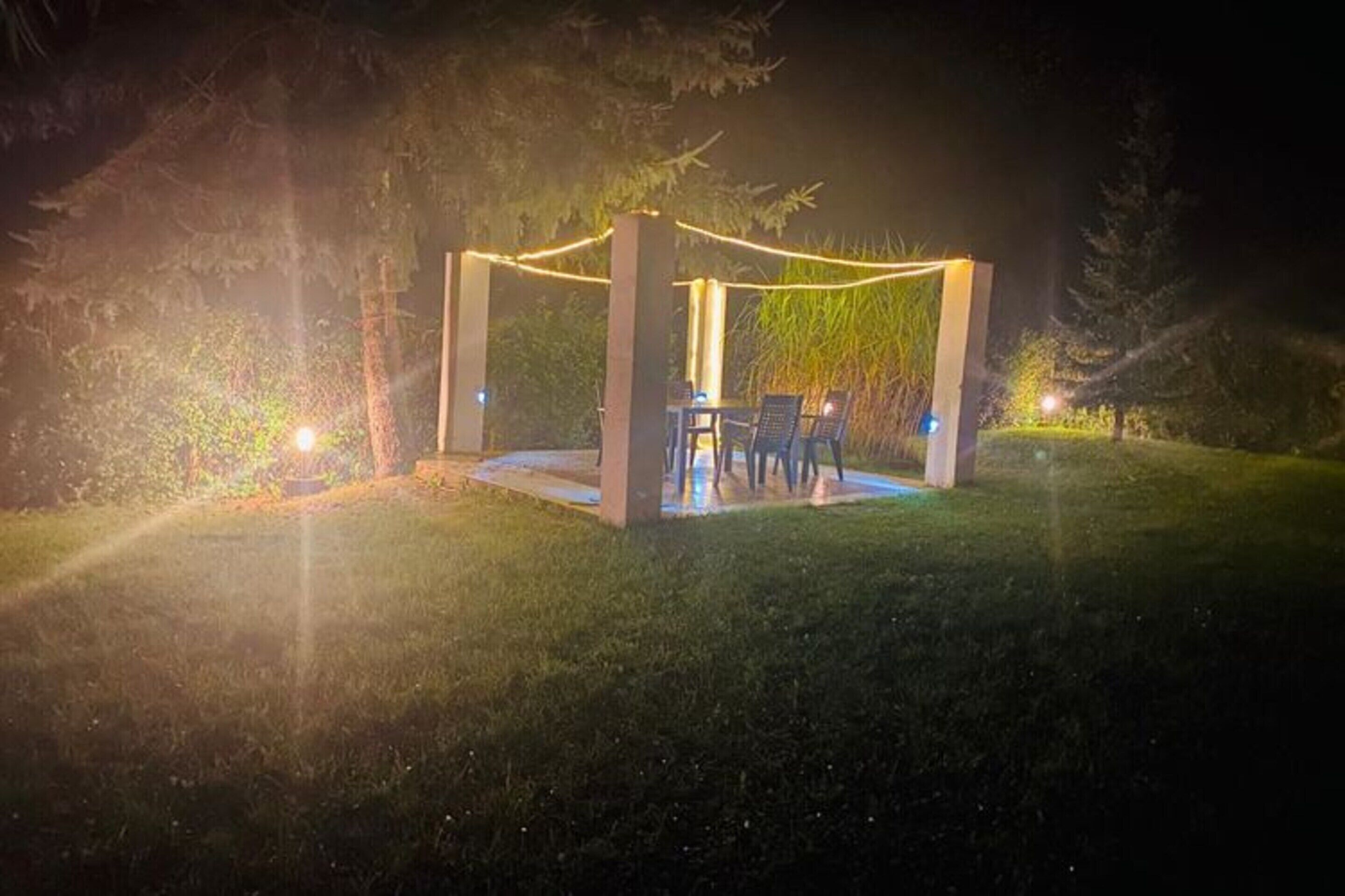 An outdoor gazebo with a table and chairs, surrounded by greenery and illuminated by string lights.