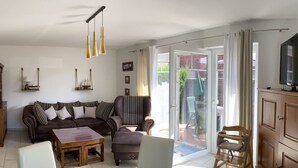 Un salon avec un canapé marron, une table basse en bois et une table à manger avec des chaises. Une porte-fenêtre coulissante donne accès à un espace extérieur équipé d’une table et de chaises.