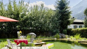 A wooden deck with a table and chairs, a small pond, and a mountain backdrop. - Maximilian Apartmenthaus 2-6 People (Neukirchen am Grossvenediger)