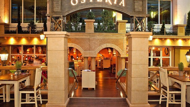 A hotel lobby with an arched entrance labeled "OSTERIA," featuring a central staircase, seating areas, and a view of the outdoor area. - Appartements im Freizeitpark Weissenhauser Strand (Wangels)