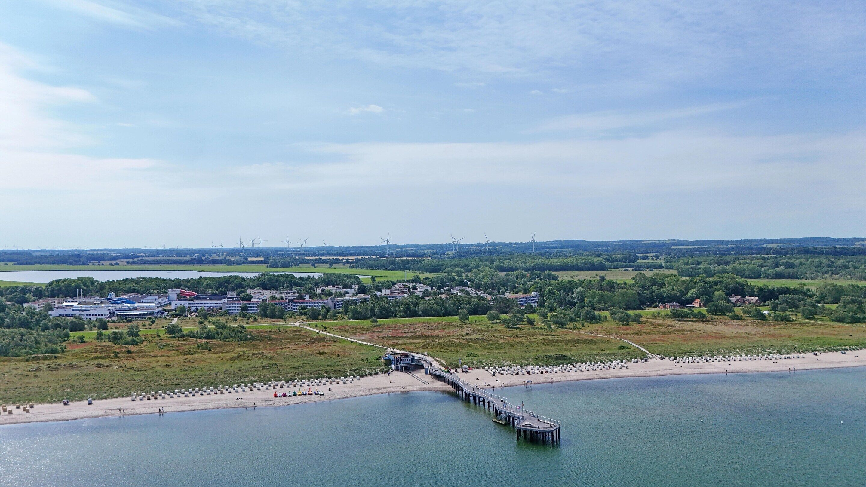Eine Küstenlandschaft mit einem Steg, der ins Wasser ragt, einem Strand voller Menschen und Windkraftanlagen in der Ferne.