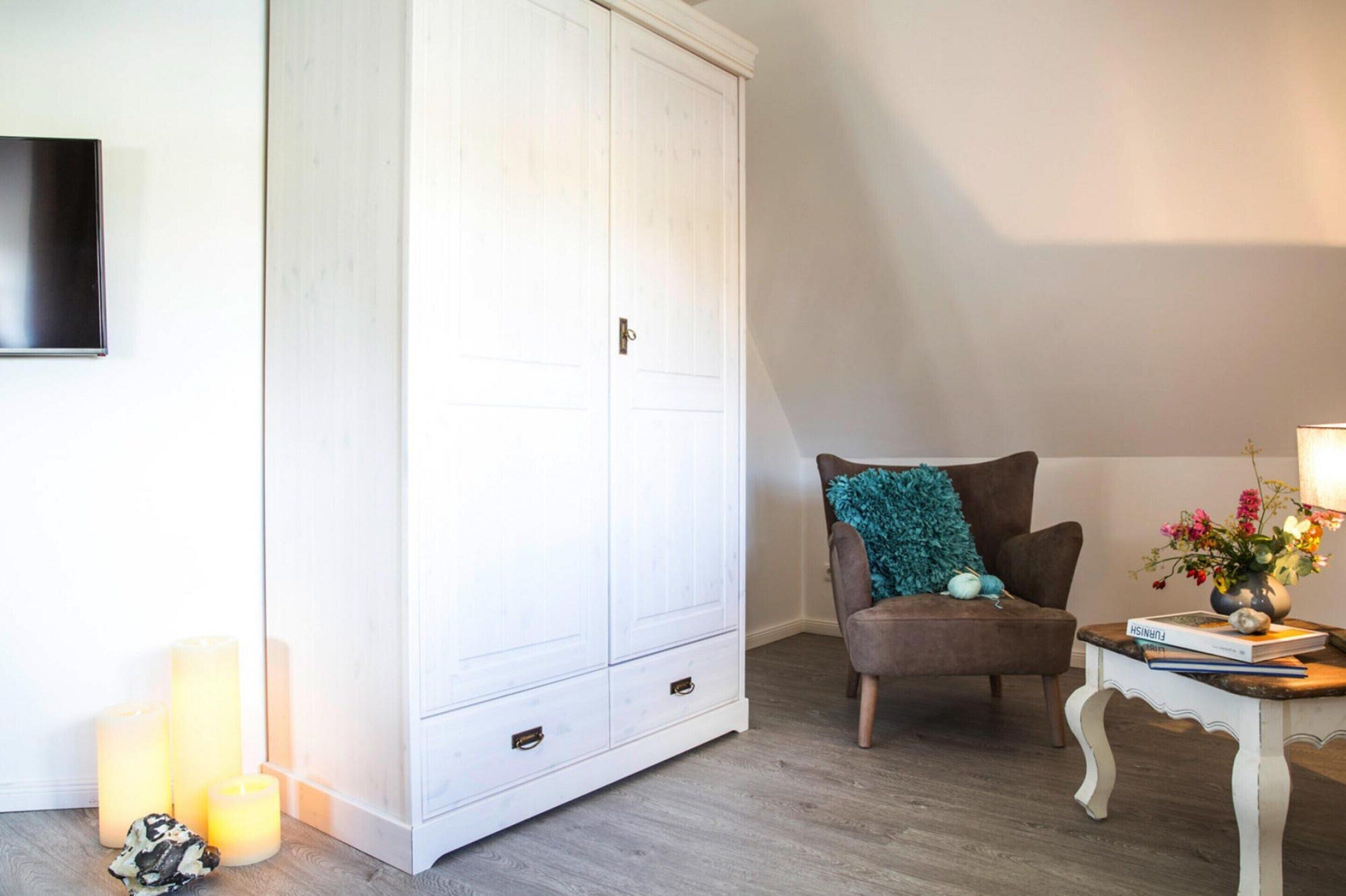 A room with a white wardrobe, a brown armchair, a small table with a lamp and flowers, and a TV mounted on the wall.