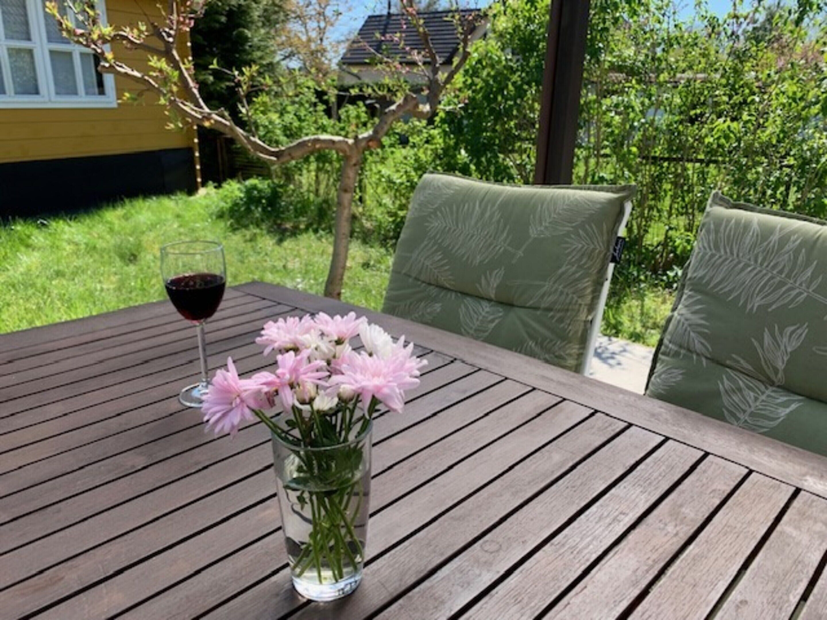 A wooden table with a glass vase containing pink flowers and a glass of red wine.