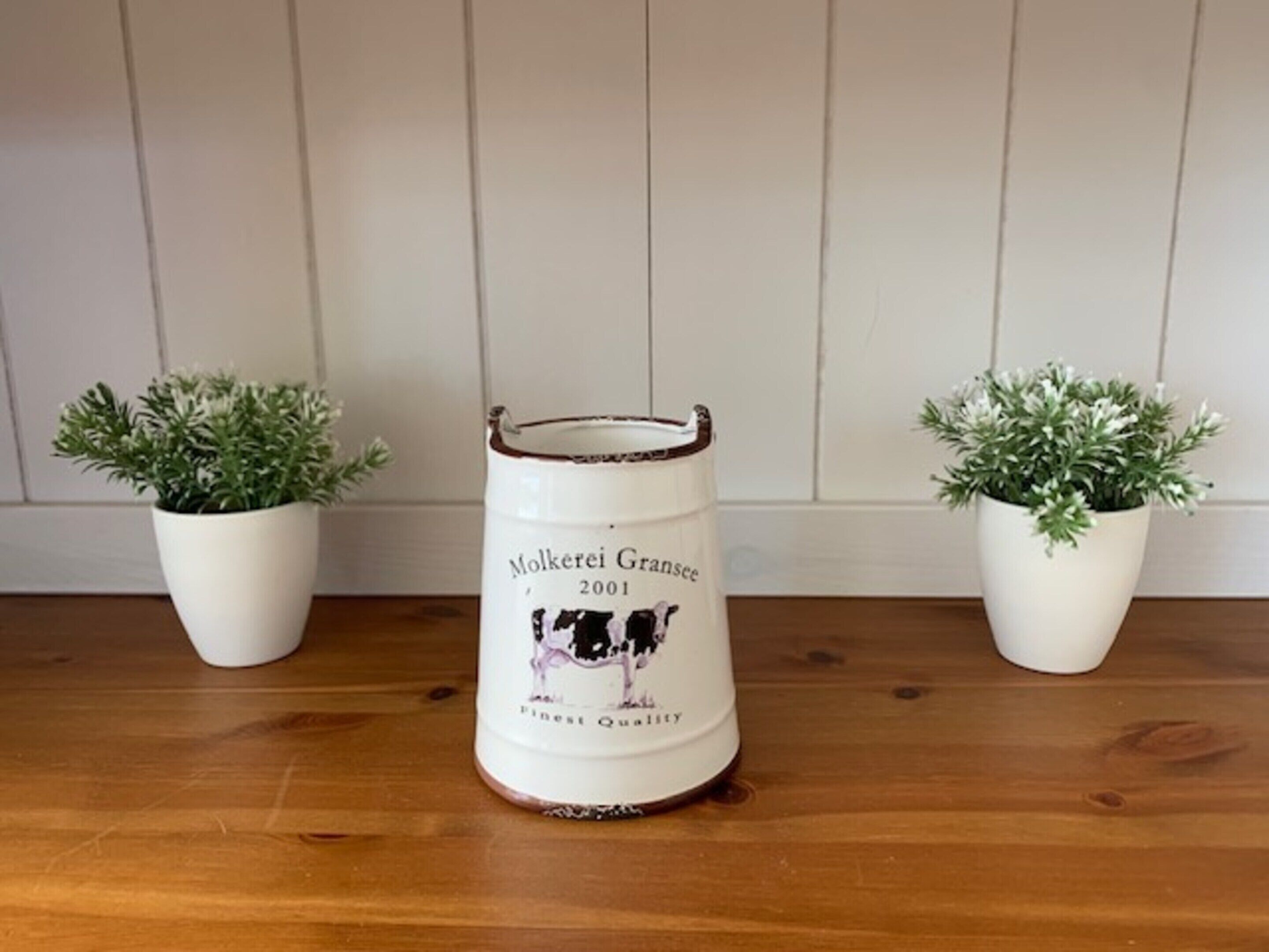 A white ceramic container with a cow illustration and text, placed on a wooden surface with two potted plants on either side.