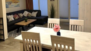 A living room with a sofa, a coffee table, and a dining table with chairs. There is a painting on the wall and a window with blinds. - Doppelhaushalfte Dunenrose, Glowe (Glowe)