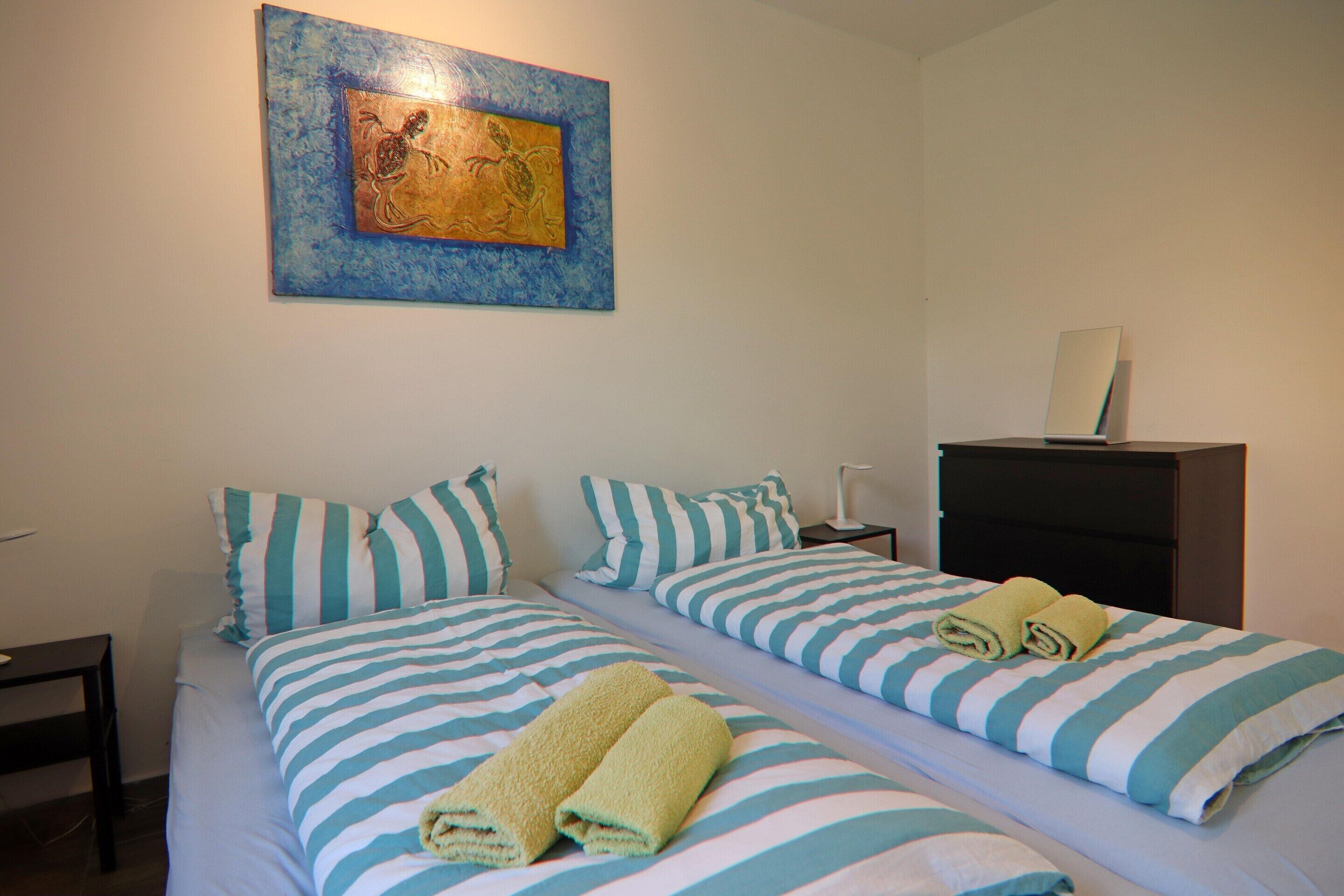 A double bed with striped bedding and two rolled towels, a nightstand, a small table, and a painting on the wall.