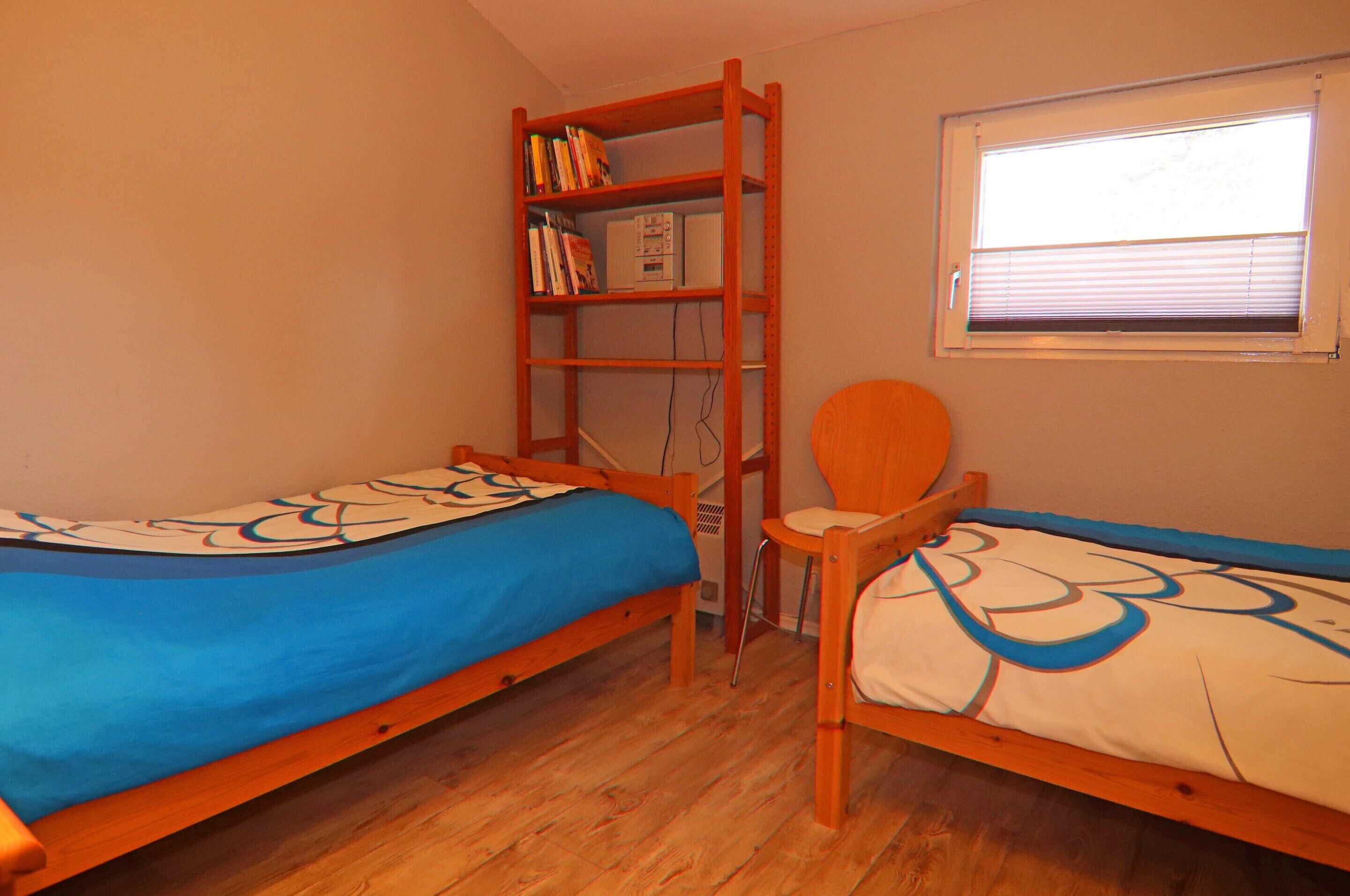 Una habitación pequeña con dos camas de madera, una estantería, una silla y una ventana con persianas.
