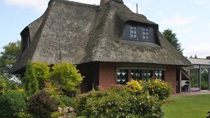 Un charmant cottage au toit de chaume, avec une base en briques, entouré de jardins bien entretenus et sous un ciel dégagé.