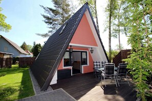 Een driekantig huis met een donker dak en een rode gevel, met een terras en tuinmeubilair en een glazen schuifdeur als ingang.