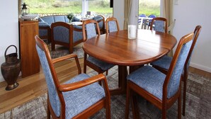 A dining room with a round wooden table, six blue upholstered chairs, a white candle, and a view of the outdoors through large windows. - Ferienhaus am Watt, List auf Sylt (List)