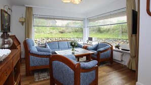 A cozy living room with a sofa set, a coffee table, and a TV. There are large windows with a view of greenery outside. - Ferienhaus am Watt, List auf Sylt (List)