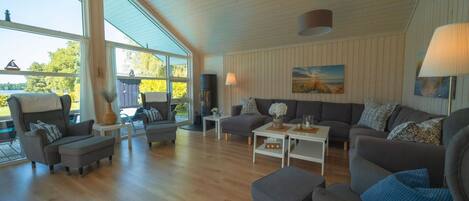 En rummelig stue med højt til loftet, store vinduer og et hyggeligt opholdsområde med sofa og lænestole.