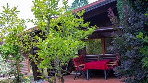 En træhytte med en overdækket terrasse, et spisebord med stole og et lille træ i forgrunden.