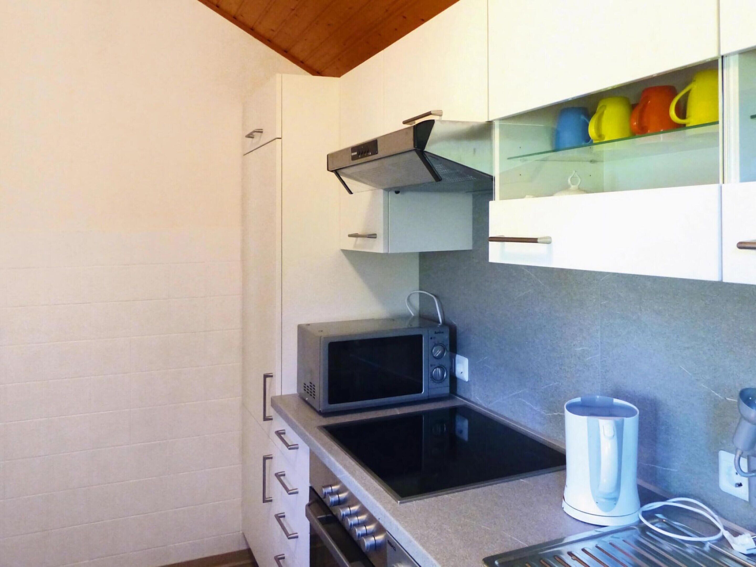 A compact kitchenette with a microwave, stove, and a glass shelf with colorful mugs.