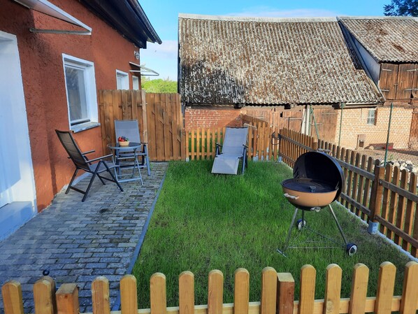 Ein Garten mit Pflasterterrasse, Holzzaun, Grill und zwei Liegestühlen.