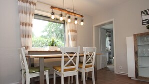 A dining area with a wooden table, four white chairs, and a hanging light fixture. - Charmantes Ferienhaus mit Kamin in Lintig (Geestland)