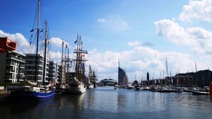 A marina with multiple sailboats docked, a modern building with balconies, and a clear blue sky. - Charmantes Ferienhaus mit Kamin in Lintig (Geestland)