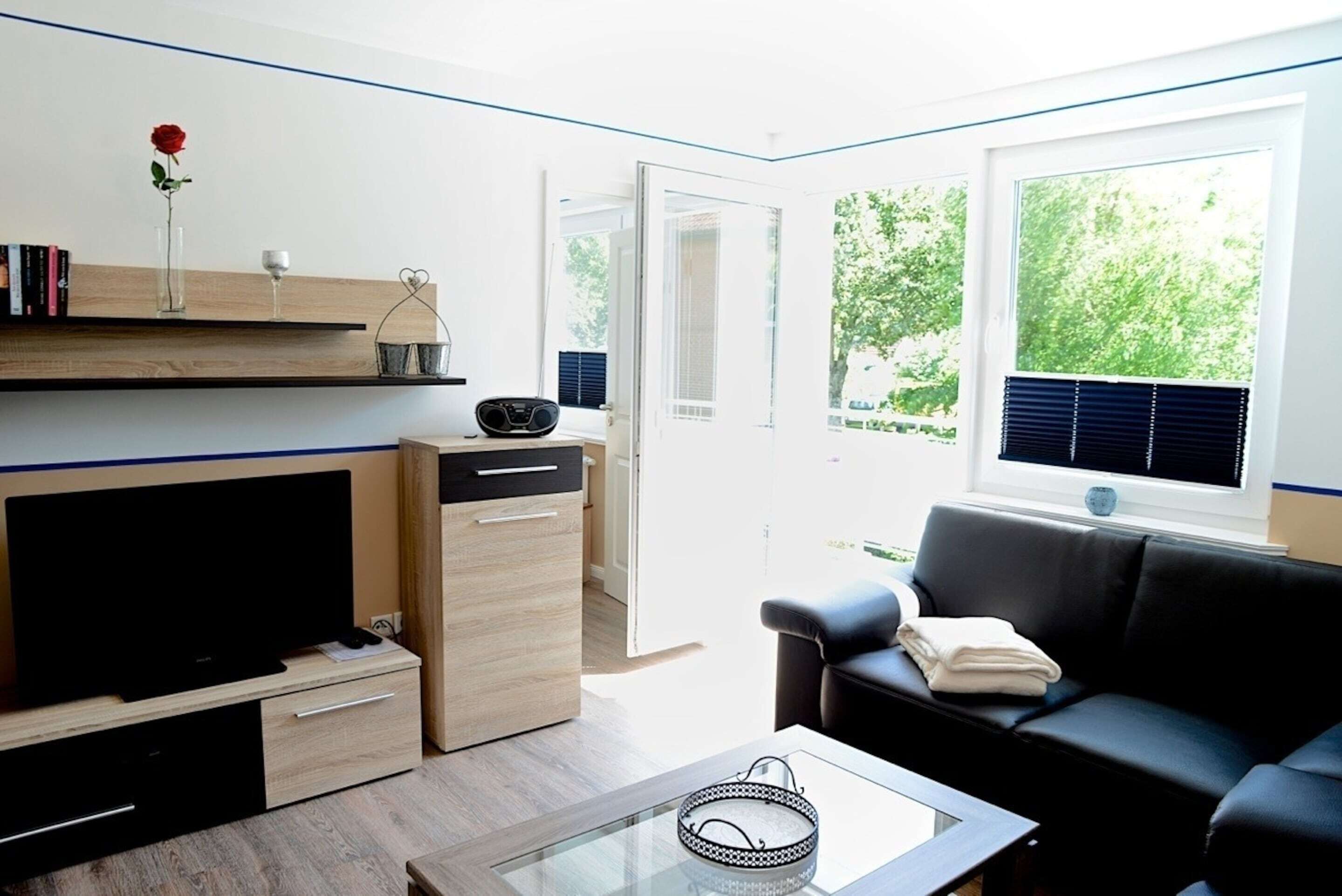 Ein modernes Wohnzimmer mit Flachbildfernseher, Couchtisch, schwarzem Ledersofa, Bücherregal und einem Fenster mit Blick auf Grünflächen.