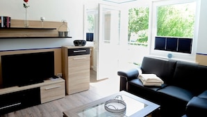 A modern living room with a flat-screen TV, a coffee table, a black leather sofa, a bookshelf, and a window with a view of greenery. - Ferienwohnung in Wyk auf Foehr (Wyk auf Foehr)