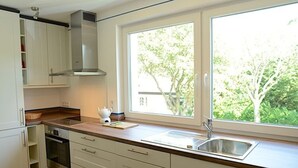 A modern kitchen with white cabinets, a stainless steel sink, and a window providing a view of trees. - Ferienwohnung in Wyk auf Foehr (Wyk auf Foehr)