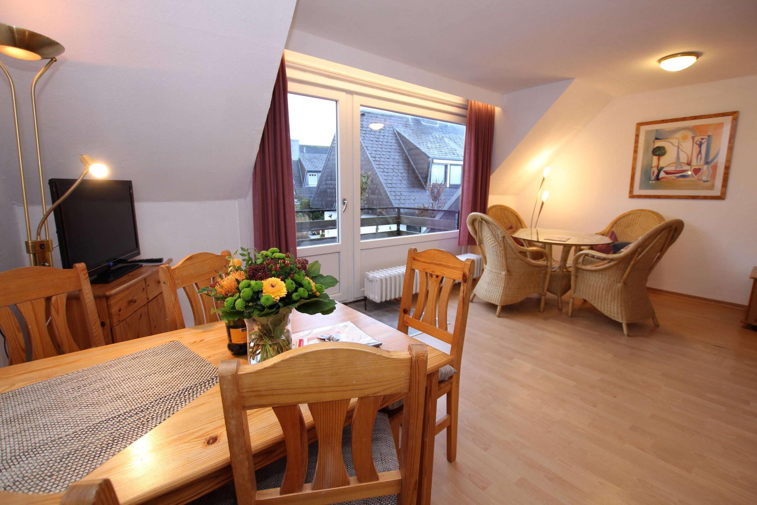 A dining area with a wooden table, chairs, a vase with flowers, and a television on a cabinet.