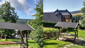 A wooden bench with a metal roof in a grassy area with a house and mountains in the background. - Ferienhaus, Oberwiesenthal (Oberwiesenthal)