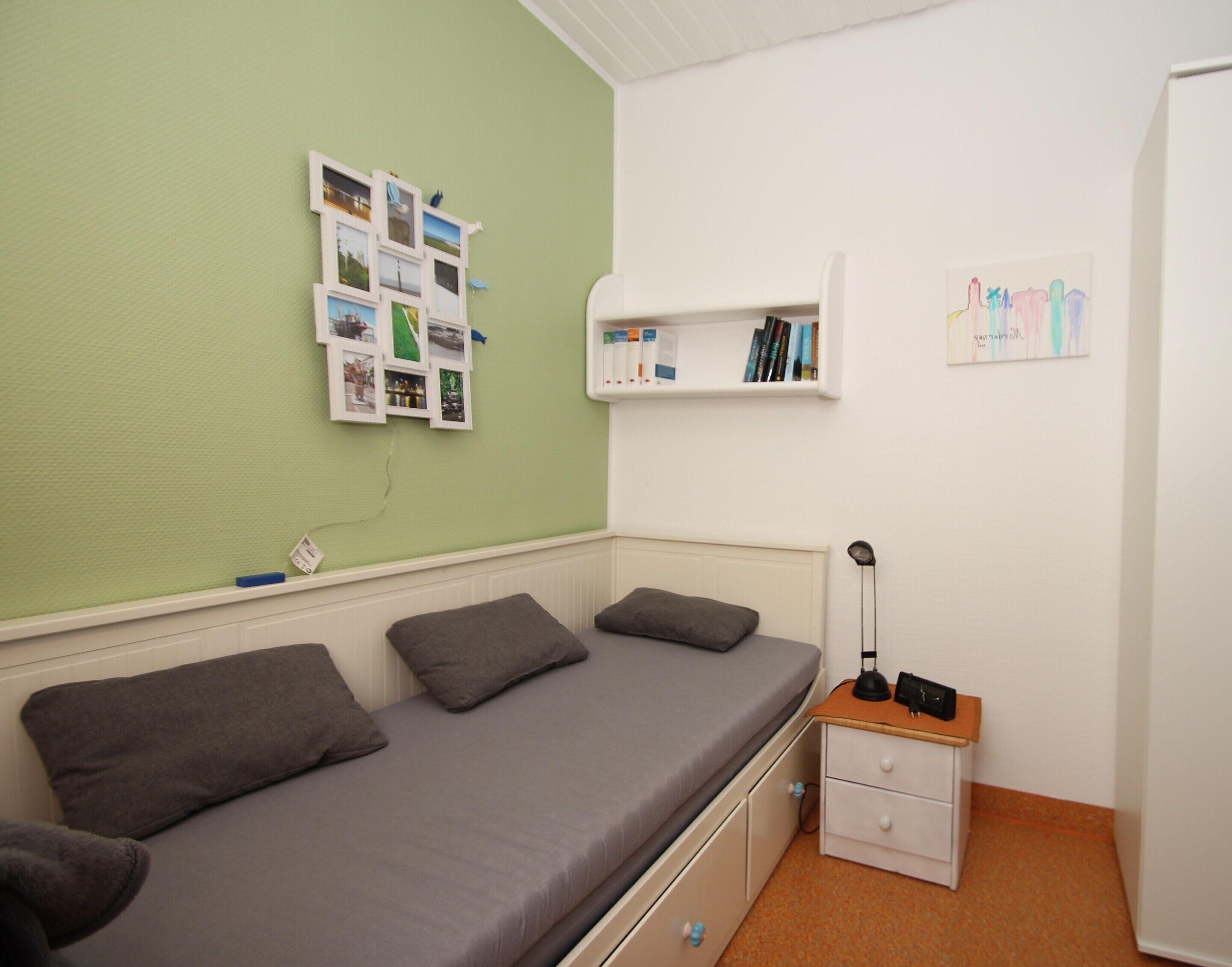 Ein Zimmer mit einem Einzelbett, einer grünen Wand, einem Nachttisch mit Lampe sowie einem Regal mit Büchern und einem Bild.