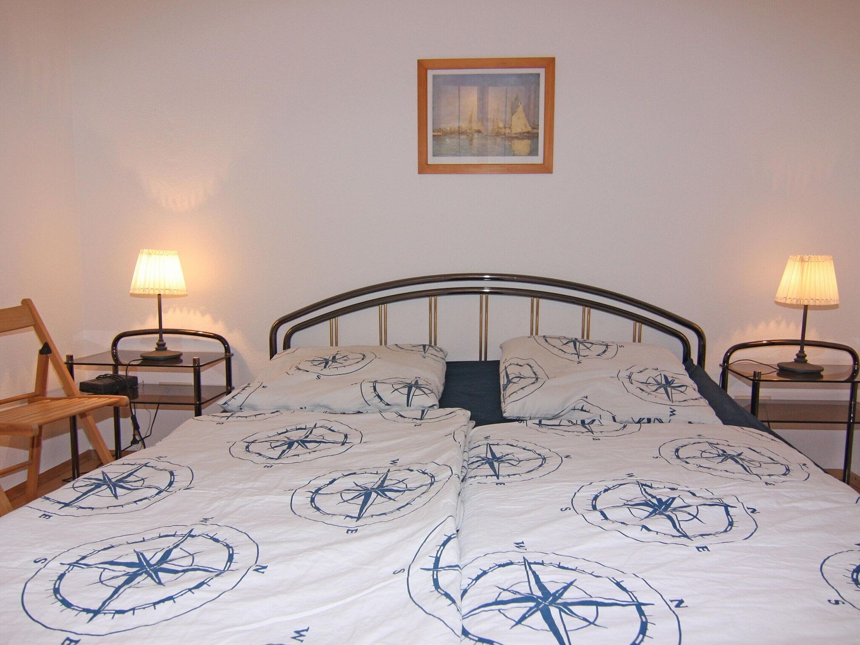 A neatly made bed with a compass-themed duvet, two bedside lamps, a wooden chair, and a framed painting on the wall.