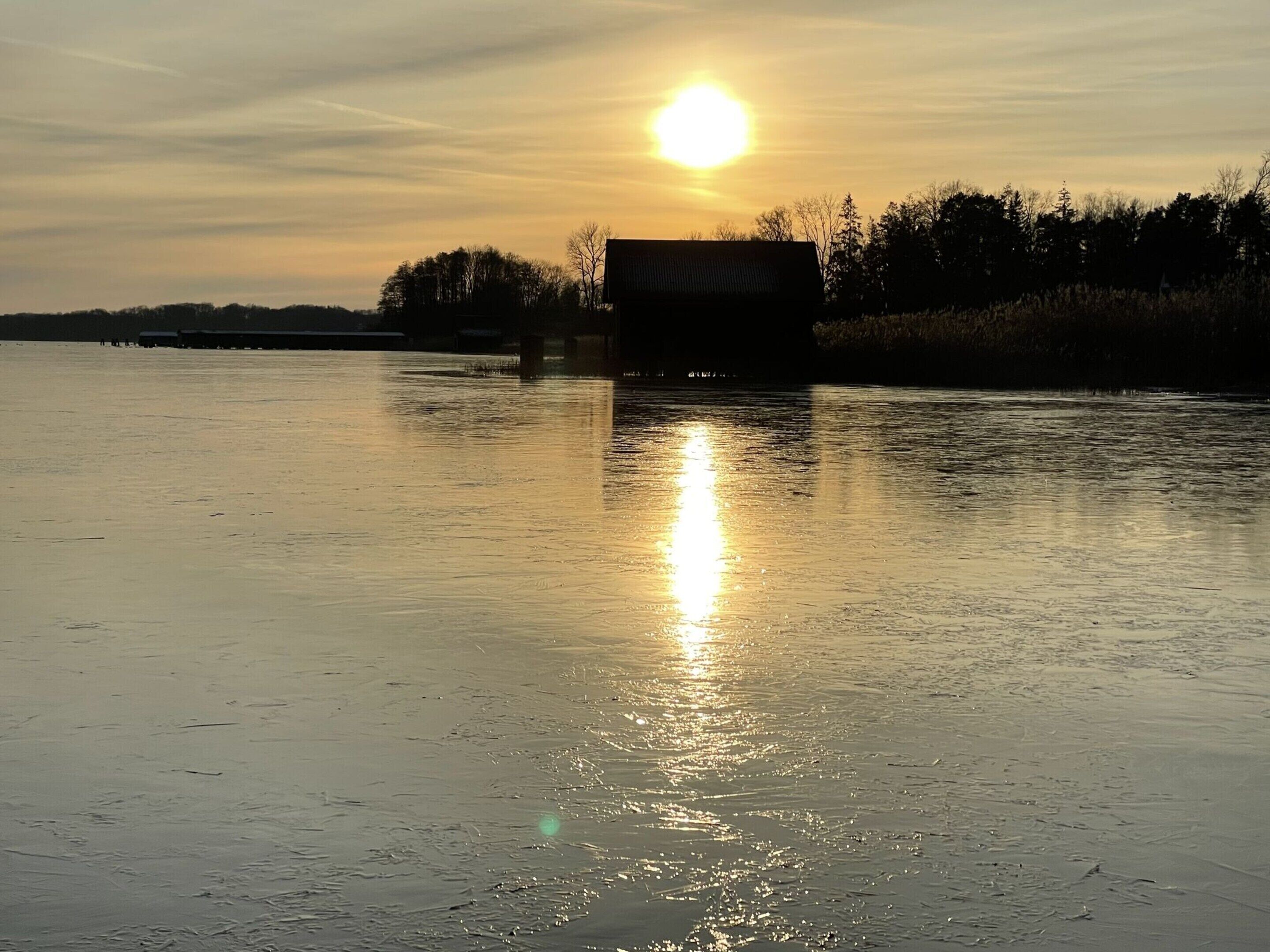 A calm body of water reflecting the sun, with a building and trees in the background.