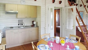 A cozy kitchen with wooden cabinets, a sink, and a clock on the wall. There is a dining area with a table set for four, featuring blue and white plates and cups. A basket of lemons is on the table, and a staircase leads to an upper level. - Ferienhaus in Lissendorf bei Eifel Wegen (Lissendorf)