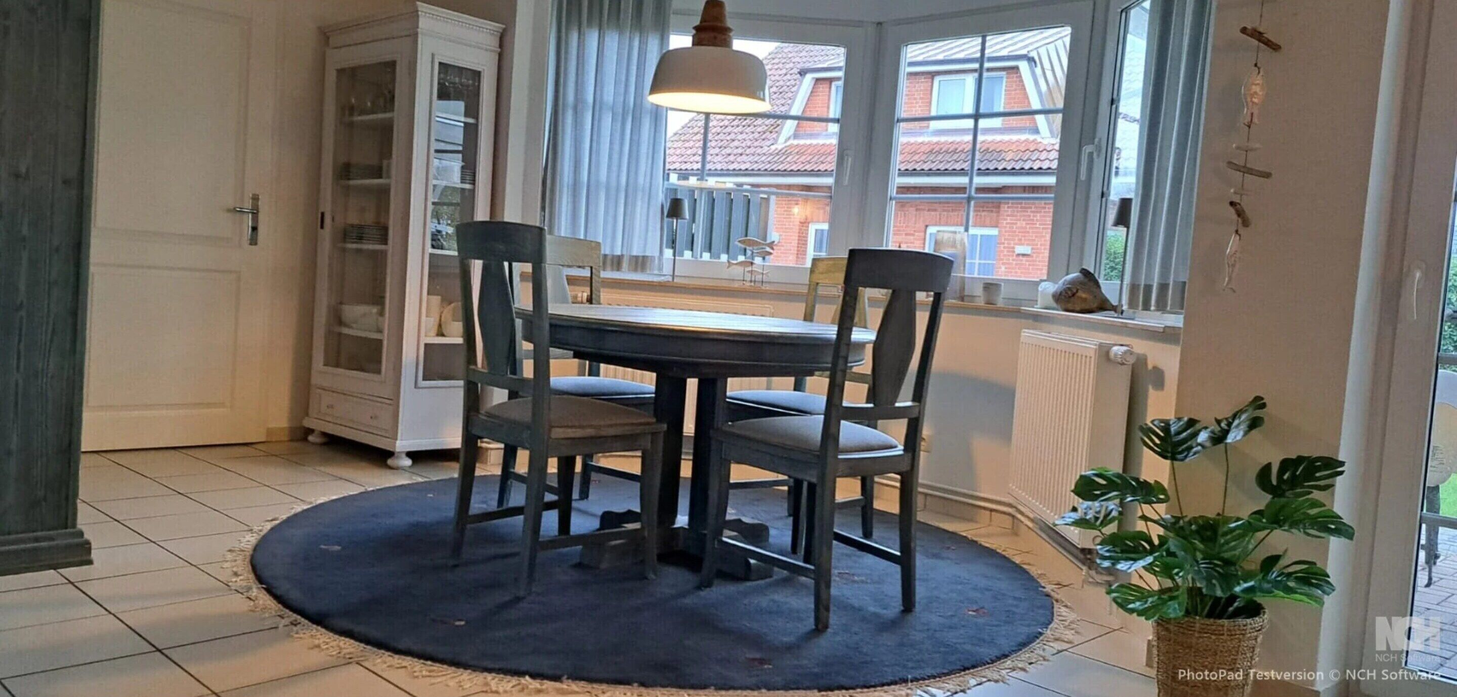 Ein Essbereich mit einem runden Tisch, vier Stühlen, einer Pendelleuchte, einem Blumentopf und Blick auf ein rotbackenfass Gebäude durch große Fenster.