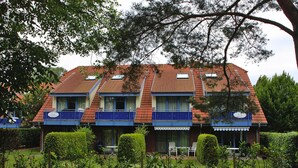 Ein zweistöckiges Gebäude mit rotem Ziegeldach, blauen Balkonen und einem kleinen Garten mit ordentlich geschnittenen Büschen und Rasen.