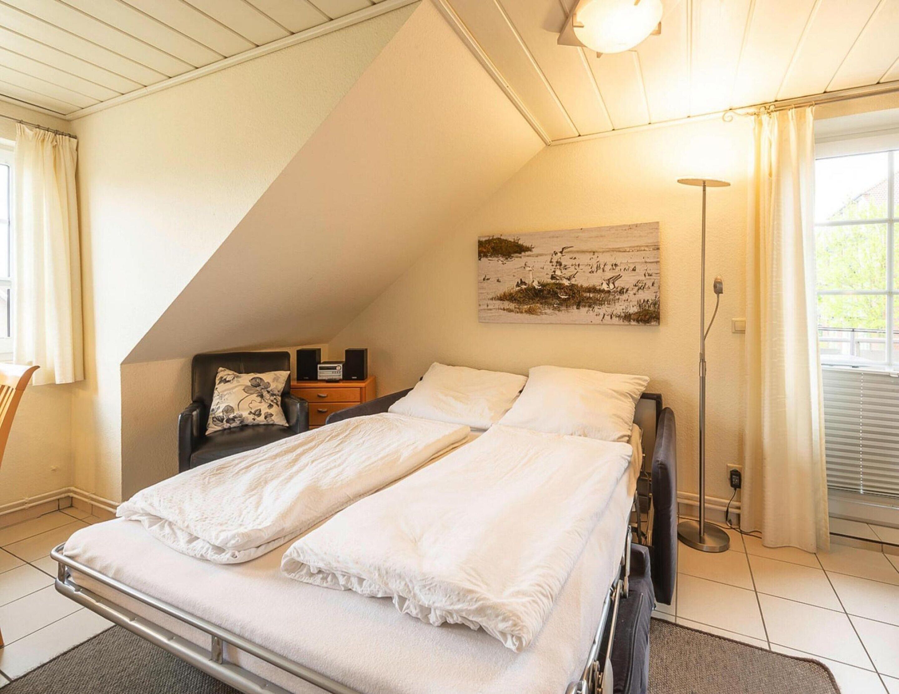 A bedroom with a sloped ceiling, a bed with white linens, a nightstand with a radio, a chair with a floral pillow, and a painting on the wall.