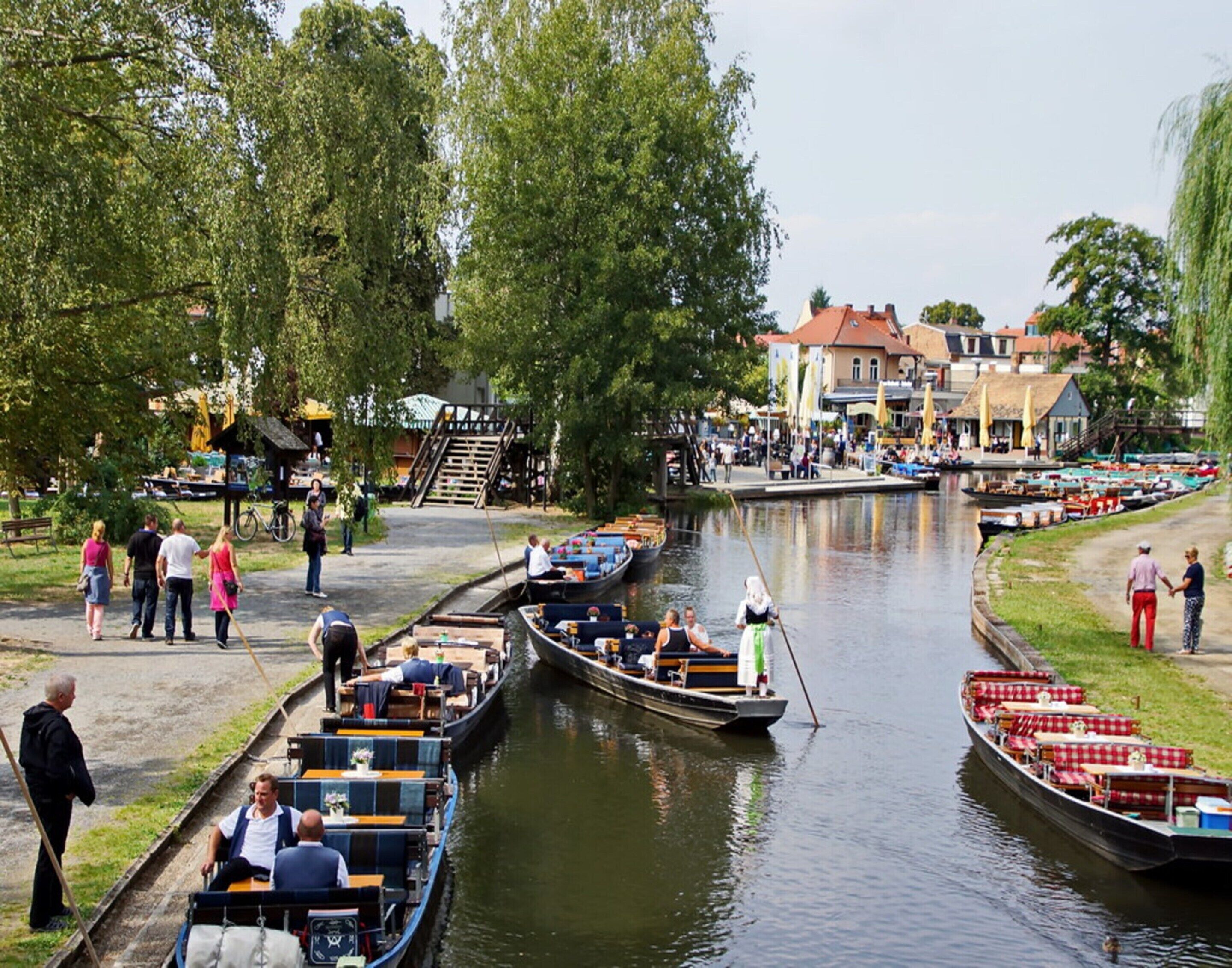 Ein Kanal mit vertäuten Booten, einem Fußweg und Gebäuden im Hintergrund.