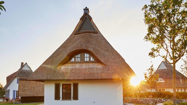Ein Haus mit strohgedecktem Dach und weißer Fassade, eine Steinmauer im Vordergrund und ein Sonnenuntergang im Hintergrund.