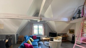 A spacious loft-style living room with a high ceiling, a large window with curtains, a blue sofa with colorful cushions, a small table, a TV, a pink ladder, and a chair. - Ferienhaus, Plau am See (Plau am See)