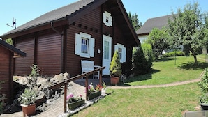 Une maison en bois avec un toit en pente, une véranda avec un banc et un jardin agrémenté de plantes en pot.