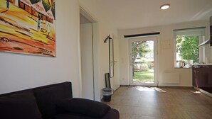 A modern living room with a dark sofa, a painting on the wall, and a view into the kitchen through an open door.