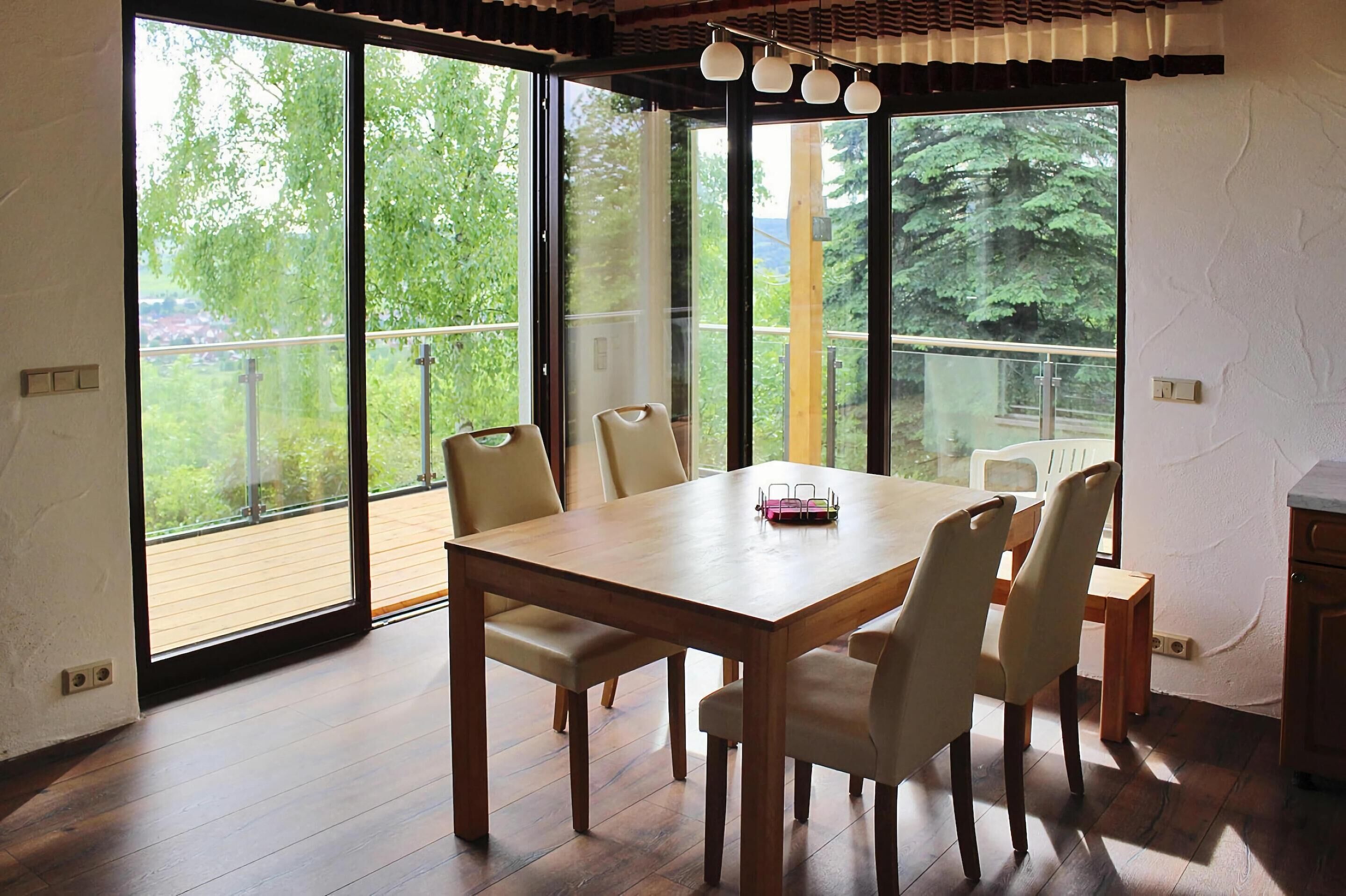 Zona pranzo con tavolo e sedie in legno, ampie finestre con vista sul verde e un balcone.