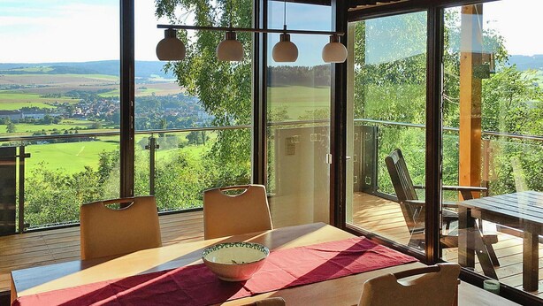 Un comedor con mesa y sillas, un mantel rojo y vistas a un paisaje verde a través de ventanas amplias.