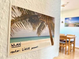 Ein an der Wand befestigtes Schild mit einer tropischen Strandansicht und dem Hinweis auf einen WLAN-Router namens 'LINUS' inklusive Passwort.