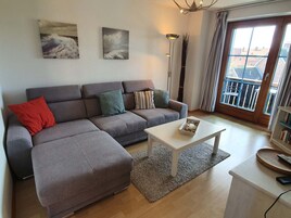 Ein modernes Wohnzimmer mit einer grauen Eckcouch, einem weißen Couchtisch und einem großen Fenster mit Blick auf Gebäude.