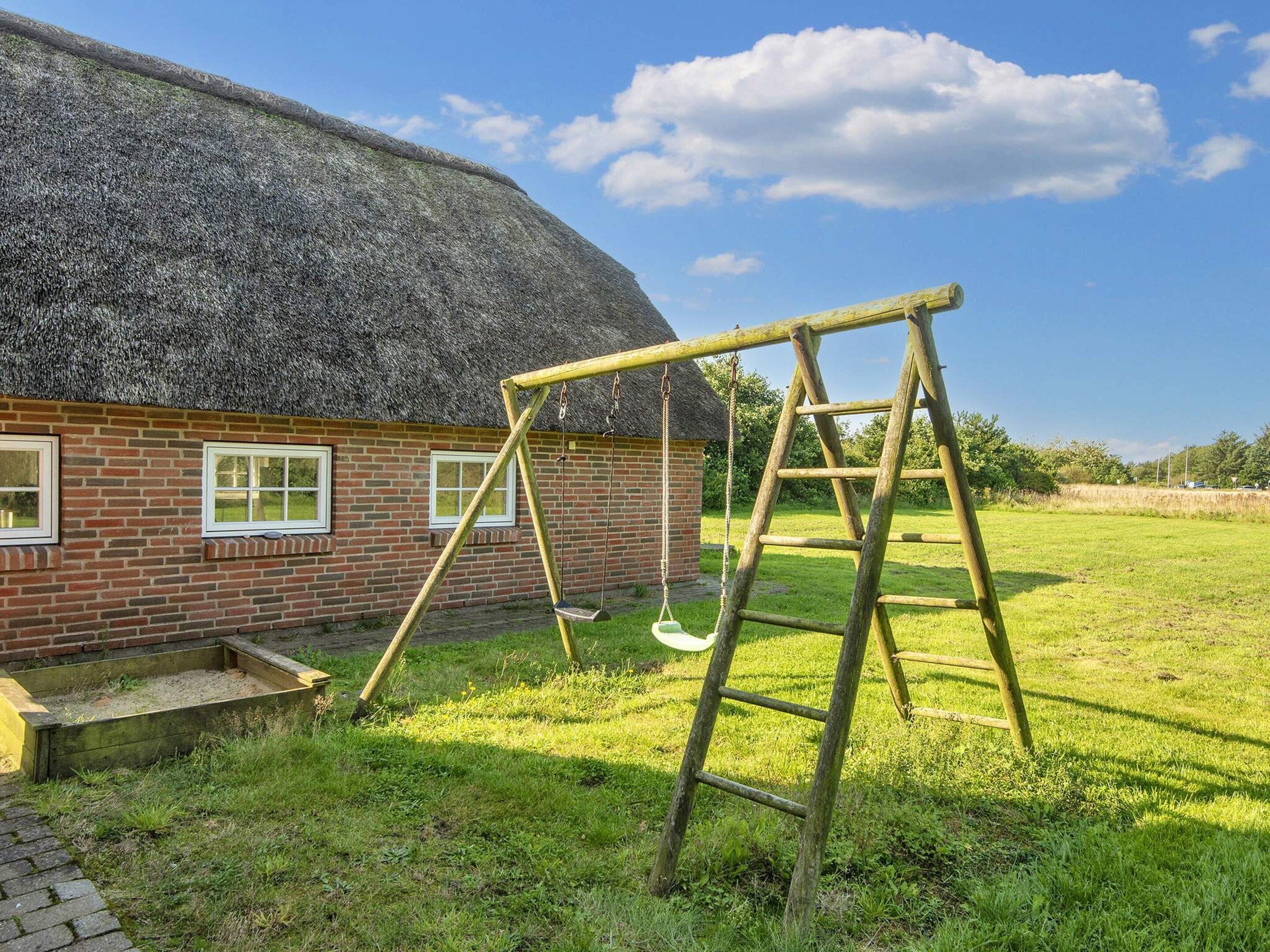 A thatched-roof house with a brick exterior, a wooden swing set, and a ladder in a grassy yard.