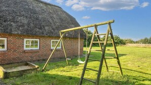 Une maison à toit de chaume, aux murs en briques, avec une balançoire en bois et une échelle dans une cour verdoyante.