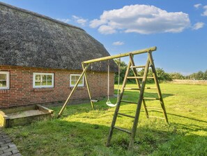 Een rieten huis met een bakstenen gevel, een houten schommel en een ladder in een gazon.