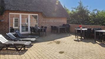 Eine Terrasse mit Liegestühlen und Tischen, ein Backsteinhaus mit einem Strohdach und Blick auf Bäume.