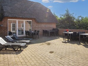 A patio area with lounge chairs and tables, a brick building with a thatched roof, and a view of trees. - 4 Star Holiday Home in Ulfborg (Ulfborg)