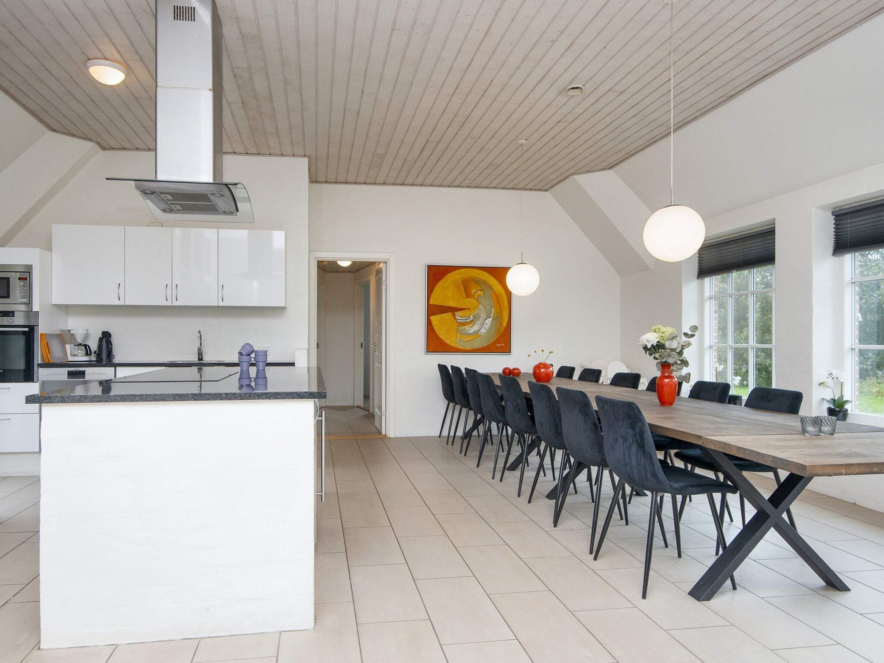 A modern kitchen with white cabinets and a central island, a dining area with a long table and chairs, and a large window providing natural light.