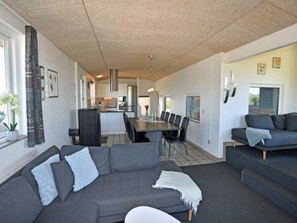 A modern living room with a grey sectional sofa, a dining table with chairs, and a kitchen in the background. - 4 Star Holiday Home in Ringkobing (Ringkobing)