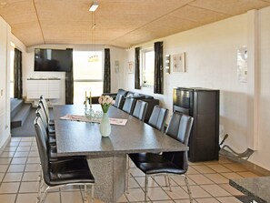Un espace repas avec une longue table, des chaises, un vase de fleurs, un réfrigérateur et, au fond, une cuisine.